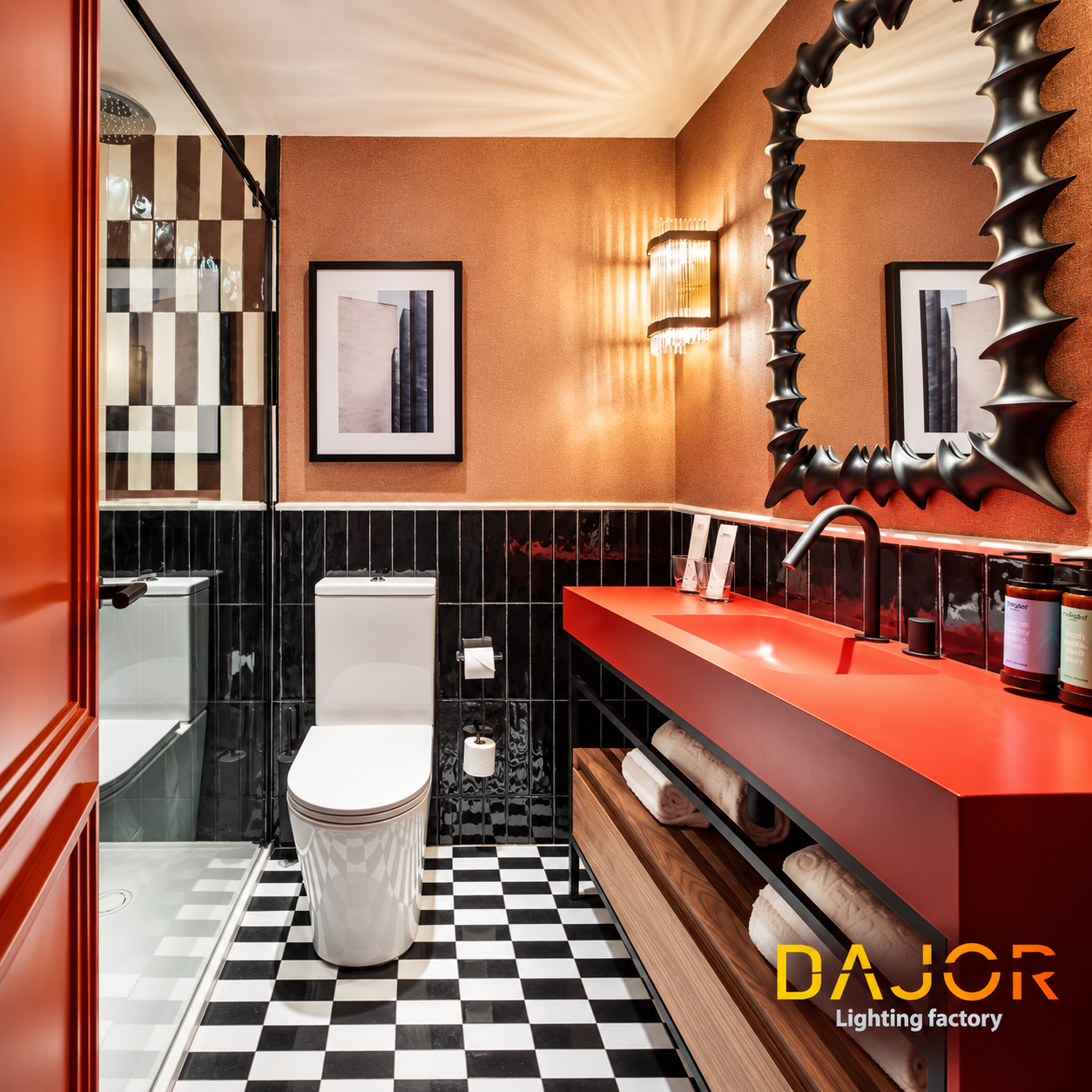 Baño de hotel con lavabo rojo, revestimientos negros y terracota, espejo escultórico y aplique decorativo de cristal.